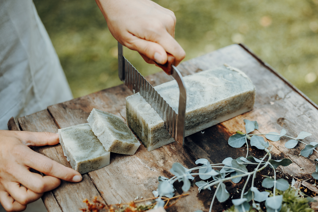 Fabrication de savon artisanal : une main coupant une barre de savon naturel à base d'herbes, illustrant le processus fait main et l'importance des ingrédients naturels dans la création de savons artisanaux.