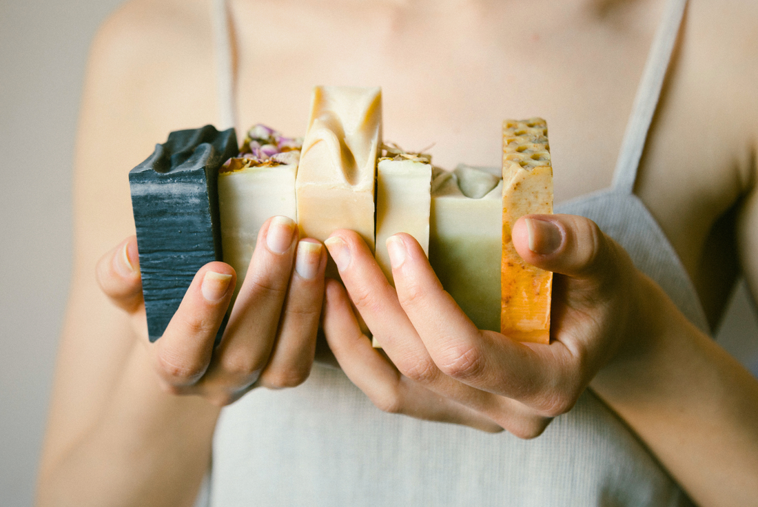 Mains tenant différents savons artisanaux de couleurs variées, mettant en avant l'importance de choisir le bon savon en fonction de votre type de peau pour un nettoyage doux et hydratant.